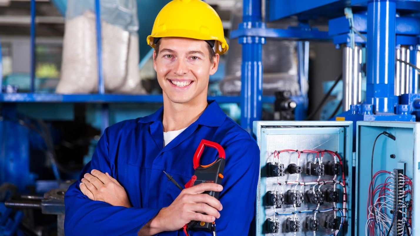 A commercial electrician in a blue jumpsuit stands next to his work.